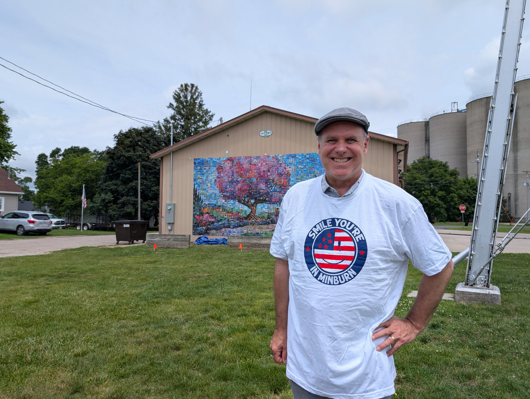 Smiling man in a white “Smile You’re In Minburn” T-shirt and cap stands on grass; mural and storage tanks behind him.